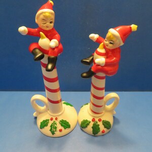 Könnte beinhalten: Zwei Kerzenhalter für Weihnachten aus Keramik. Jeder hat eine rot-weiß gestreifte Zuckerstange mit einer kleinen Elfenfigur in rotem Anzug und Hut. Der Sockel ist weiß mit grünem Stechpalmenlaub und roten Beeren. Der Elf hält eine gelbe Kerze.