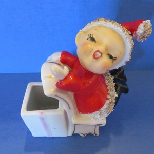 Puede incluir: Figura de cerámica vintage de un elfo sonriente con abrigo y sombrero rojos, adornada con detalles dorados y blancos. El elfo se inclina sobre una jardinera blanca con una raya rosa. Diseño retro caprichoso.
