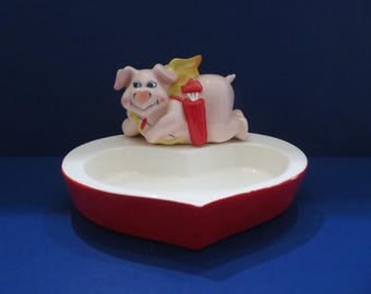 May include: A ceramic heart-shaped dish with a red exterior and white interior. A pink pig with yellow wings and a red quiver of arrows is perched on top. The pig is smiling, and the dish is on a blue surface.