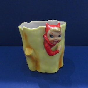 May include: A vintage ceramic planter in the shape of a tree trunk, painted yellow with brown accents. A smiling figure in a red devil costume peeks out from the side. The interior is white.