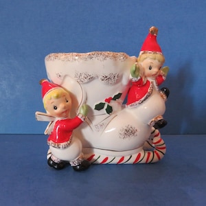 Könnte beinhalten: Ein weihnachtlicher Stiefel-Blumentopf aus Keramik mit zwei Elfenfiguren. Der Stiefel ist weiß mit goldenen Akzenten und einem rot-grün gestreiften Sockel. Die Elfen tragen rote Mäntel und Hüte.