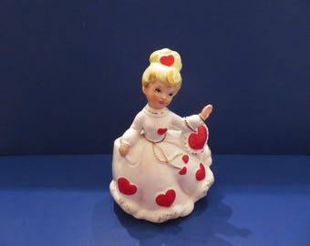 May include: A ceramic figurine of a young woman in a white dress adorned with red hearts. The figurine has blonde hair and is holding a heart-shaped accessory. Gold accents are present on the dress and around the base. The background is a solid blue.