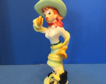 May include: A vintage ceramic cowgirl figurine. She wears a white top, green skirt, and black boots. The figurine has a light green hat, a red scarf, and yellow gloves, holding a yellow lasso. The style is retro and cartoonish.