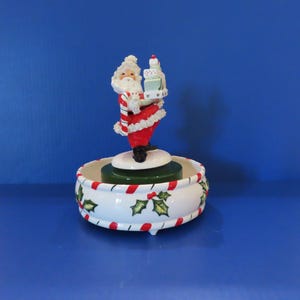 May include: A ceramic musical figurine featuring a Santa Claus figure holding a stack of gifts and a candy cane, standing atop a white circular base decorated with green holly leaves and red berries, accented by red and white striped trim.