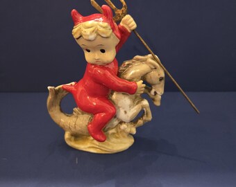 May include: A vintage figurine depicting a small devil in a red outfit with horns, riding a seahorse. The devil figure holds a gold trident. The seahorse is cream-colored with a detailed, textured design.