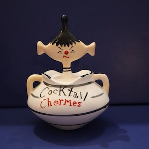 Op de afbeelding: Een vintage keramische cocktail kers pot met een grillig gezichtsontwerp. De pot is wit met zwarte en rode letters die "Cocktail Cherries" luidt. Het heeft aan weerszijden een handvat en een decoratieve kop erboven.