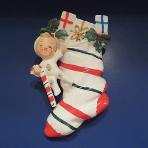 Puede incluir: Una media de Navidad de cerámica blanca con rayas rojas y verdes. Una pequeña figurita de cerámica de un niño que sostiene un bastón de caramelo está unida a la media.