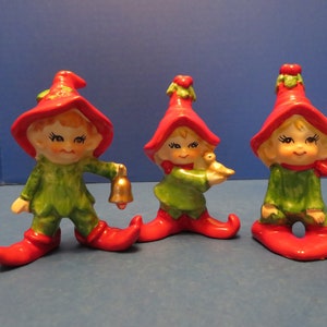 Puede incluir: Tres figuras de elfos de cerámica. Cada elfo lleva un sombrero y zapatos rojos, una túnica verde y tiene una cara sonriente. Un elfo sostiene una campana dorada, otro sostiene un pajarito y el tercero tiene un pájaro posado en su hombro. Las figuras son de temática navideña.