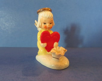 May include: A porcelain figurine of a kneeling angel holding a red heart. The angel is wearing a yellow dress and a gold halo. The figurine is sitting on a white cloud base.