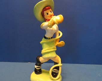 May include: A ceramic figurine of a cowgirl in a green hat and skirt, white top, and black boots. The cowgirl is holding a yellow lasso. The figurine is on a yellow base.