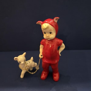 May include: A vintage ceramic figurine of a child in a red outfit with a matching hood, holding a leash attached to a small white dog with black spots. The dog has a gold-colored chain collar. The child has blonde hair and is standing on a blue surface.