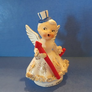 Könnte beinhalten: Weiße Porzellanfigur eines Engels, der einen blau-weißen Hut trägt und einen roten Pfeil hält. Die Basis der Figur trägt die Aufschrift "JULY ANGEL".