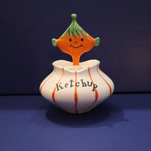 Pode incluir: Um dispensador de ketchup de cerâmica com um design fantasioso. O recipiente branco tem listras verticais laranja e a palavra "Ketchup" em preto. Uma figura laranja sorridente com cabelo verde está no topo do dispensador.