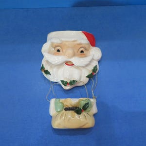 Puede incluir: Un bolsillo de pared de cerámica de Papá Noel. La cara de Papá Noel tiene barba blanca y gorro rojo. La bolsa es beige con detalles verdes. El fondo es azul.