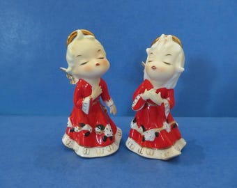 May include: Two ceramic figurines of angels, each with closed eyes, wearing red dresses with white and black accents. They have gold halos and wings, with a cream-colored face and hair. The figurines are on a blue background.