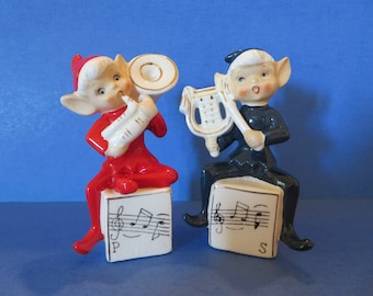 May include: A pair of vintage ceramic elf figurines. One is in red playing a wind instrument, the other in black playing a harp. Both are seated on blocks with musical notes. The figurines have white hair and pointed ears.