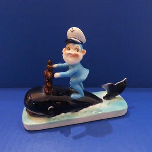 Puede incluir: Una figura de cerámica que muestra a un marinero con barba blanca y uniforme azul, con una gorra de capitán blanca con un emblema de ancla. El marinero está sobre una ballena negra brillante, situada en una base azul claro con detalles de espuma.
