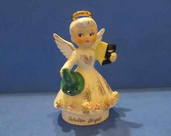 Könnte beinhalten: Keramik-Engelsfigur mit goldenem Heiligenschein, weißen Flügeln und einem weißen Kleid mit goldenen Akzenten. Der Engel hält einen grünen Hut und ein schwarz-gelbes Buch. Auf dem Sockel steht "October Angel".