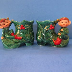 Könnte beinhalten: Zwei Keramik-Weihnachtselfen-Pflanzgefäße. Jedes Pflanzgefäß hat die Form eines grünen Stechpalmenblatt-Stiefels mit einem roten Beerencluster. Auf jedem Stiefel sitzt ein Elf in einem grünen Anzug und roten Schuhen.