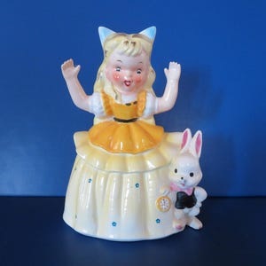 Op de afbeelding: Een porseleinen beeldje van een meisje in een gele jurk en een wit konijn. Het meisje heeft blond haar en draagt een blauwe strik. Het konijn houdt een zakhorloge vast.