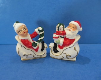 Könnte beinhalten: Zwei Weihnachtsfiguren aus Keramik. Eine ist Mrs. Claus in einem Schlitten, die eine Zuckerstange hält. Die andere ist der Weihnachtsmann in einem Schlitten, der verpackte Geschenke hält. Beide Figuren tragen rot-weiße Outfits mit goldenen Akzenten.