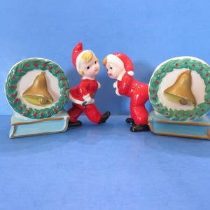 Könnte beinhalten: Zwei keramische Weihnachtsfiguren von Kindern in roten Anzügen und Hüten. Jede Figur steht in der Nähe eines dekorativen Kranzes mit einer goldenen Glocke. Die Kränze stehen auf hellblauen, buchförmigen Sockeln. Der Hintergrund ist einfarbig blau.