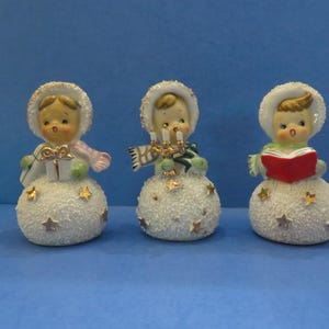 Puede incluir: Tres figuras de cerámica de muñecos de nieve con detalles de estrellas. Cada muñeco de nieve lleva un sombrero blanco y tiene un accesorio diferente: un regalo, velas y un libro. Los muñecos de nieve tienen mejillas rosadas, sobre fondo azul.