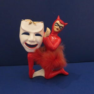 Könnte beinhalten: Eine Keramikfigur mit einer weißen Komödienmaske und einer roten Teufelsfigur mit Federrock. Die Teufelsfigur kniet, mit einer schwarzen Maske und Hörnern. Die Maske hat einen goldenen Rand und lächelt. Die Figur steht auf einer blauen Oberfläche.