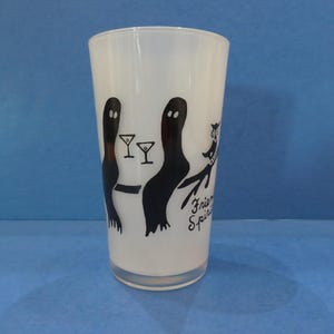 May include: White glass with a black graphic of two ghost-like figures, martini glasses, and a cat-like figure. The words "Friend Spirits" are written in a script font on the glass. The glass is cylindrical and sits on a blue surface.
