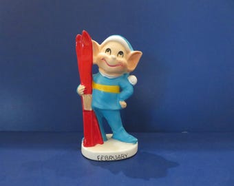 May include: A ceramic figurine of a smiling elf wearing a blue outfit with a yellow stripe and a white hat. The elf is holding red skis and stands on a base that reads "FEBRUARY". The figurine has a vintage, cartoonish style.
