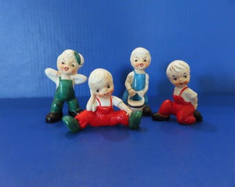 May include: Four ceramic figurines of children with blonde hair and painted facial features. One stands in green overalls, one sits in red overalls, one stands in blue overalls holding a bucket, and one sits in red overalls with black shoes.