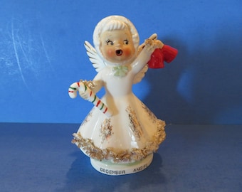 May include: A white porcelain figurine of a December angel holding a candy cane and a red stocking. The angel is wearing a white dress with gold trim and a white bonnet. The base of the figurine is green and reads "December Angel".