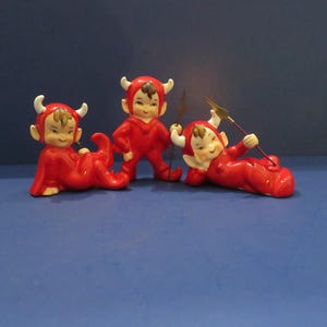 May include: Three ceramic devil figurines in red outfits with white horns and brown hair. One is standing, and two are reclining. One holds a spear with a gold tip. The figurines are on a blue surface.