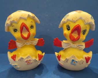 May include: A pair of yellow ceramic chick salt and pepper shakers. The chicks are wearing red and white bows and are sitting in cracked eggshells with blue and pink flowers.