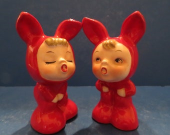 May include: A pair of red ceramic salt and pepper shakers shaped like children wearing bunny costumes. The shakers have painted facial features and are in a singing pose.