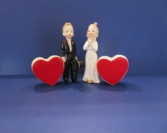 May include: Vintage wedding cake toppers featuring a bride and groom figurine, with two red heart-shaped boxes. The groom is in a black tuxedo, and the bride wears a white dress. The background is a solid blue.