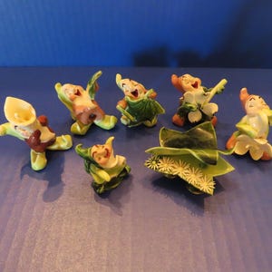 Peut inclure: Une collection de six petites figurines en céramique représentant des personnages fantaisistes interagissant avec diverses feuilles vertes et fleurs blanches. Chaque figurine présente une pose unique, avec des détails peints en vert, blanc et marron.