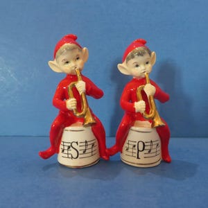 May include: Two ceramic elf figurines in red outfits and hats, playing gold trumpets. They sit on white drums with musical notes and the letters "S" and "P". The background is a solid blue.