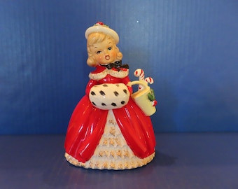 Może przedstawiać: Ceramiczna figurka przedstawiająca postać w czerwonym płaszczu z białym futrzanym kołnierzem, białym kapeluszu i długiej czerwonej spódnicy ze złotymi akcentami. Postać trzyma białą mufkę w czarne kropki oraz jasne żółte wiaderko z laskami cukrowymi.
