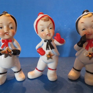 Puede incluir: Tres figuras de cerámica vintage de niños con trajes de nieve blancos con detalles rojos y negros. Cada figura sostiene una estrella dorada y tiene una pajarita roja. Las figuras tienen mejillas rosadas y están en diferentes poses.