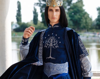 May include: A person wearing a blue velvet robe with silver embroidery and a silver and gold crown. The robe has a white tree design on the front. The person is standing in front of a body of water.