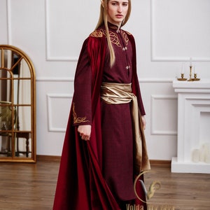 May include: A person wearing a burgundy robe with gold embroidery and a gold sash. The robe has a long, flowing red cape. The person is wearing a gold crown and has long blonde hair.