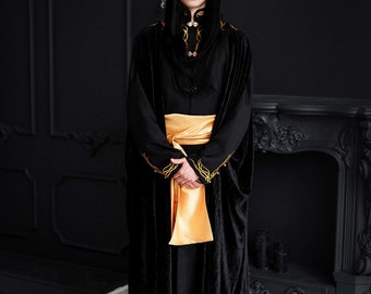 May include: A person wearing a black velvet robe with gold embroidery and a gold sash. The person is also wearing a crown with black and white accents. The background is a dark room with a fireplace.
