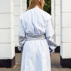 May include: A person wearing a white long-sleeved tunic with a silver sash. The tunic has a textured fabric and the sleeves are made of a smooth, silky material. The person is wearing a silver hair accessory.