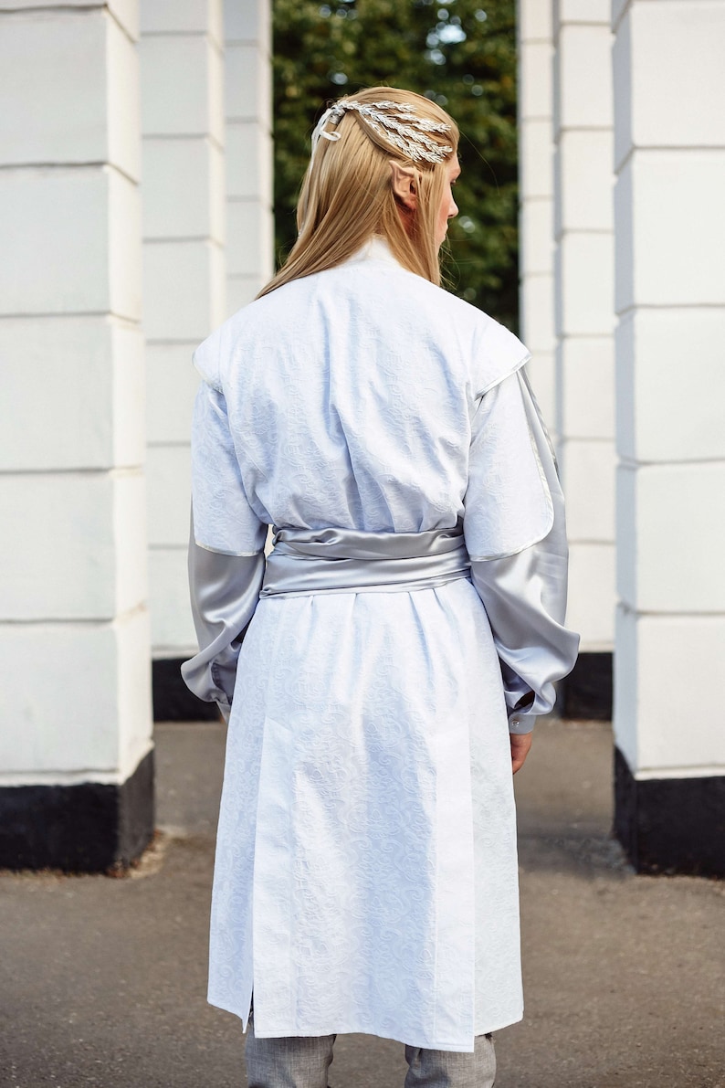 May include: A person wearing a white long-sleeved tunic with a silver sash. The tunic has a textured fabric and the sleeves are made of a smooth, silky material. The person is wearing a silver hair accessory.
