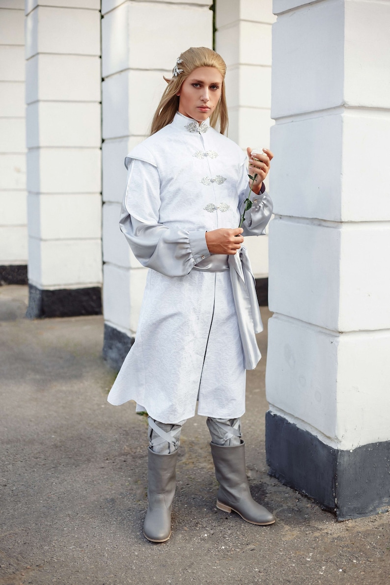 May include: A person wearing a white, long-sleeved tunic with silver buttons and a silver belt. They are holding a single white rose and wearing grey boots.
