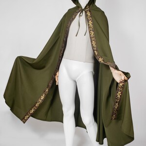 Puede incluir: Una capa con capucha de color verde oliva que presenta un borde decorativo con un patrón floral dorado a lo largo de los bordes frontales y la capucha. La capa incluye finas cintas de tela en el cuello y cubre un maniquí blanco de cuerpo entero.