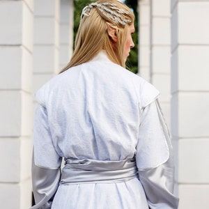 May include: A person wearing a white robe with a silver sash and a silver hairpiece. The person is standing in a white columned hallway.