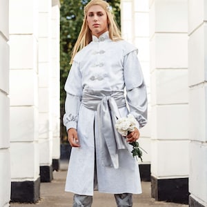 May include: A person wearing a white long-sleeved tunic with a silver sash and matching boots. They are holding a bouquet of white flowers.