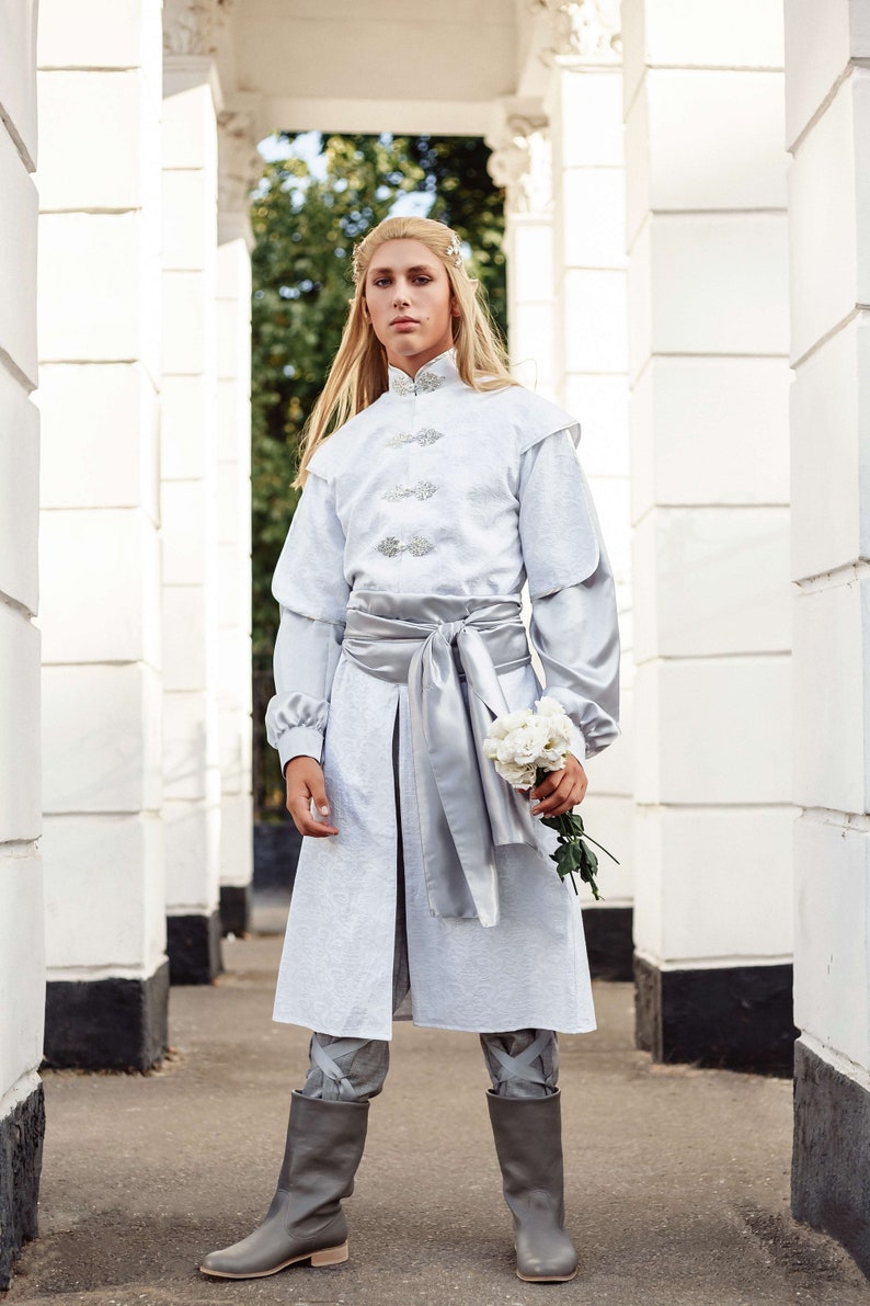 May include: A person wearing a white long-sleeved tunic with a silver sash and matching boots. They are holding a bouquet of white flowers.